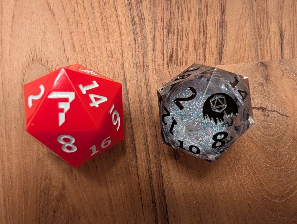 The image shows two twenty-sided dice on a wooden surface. The die on the left is red with white numerals and white Factal logo, featuring numbers like 2, 14, 19, 8, and 16 visible. Its surface is glossy and smooth. The die on the right is clear with a slightly smoky appearance, containing black numerals and sparkly inner elements. Numbers visible include 2, 12, 8, 14, and 10. This die also has a black Full Moon Storytelling logo resembling a full moon rising over trees with a hexagonal shape inside, located near the numeral 2.