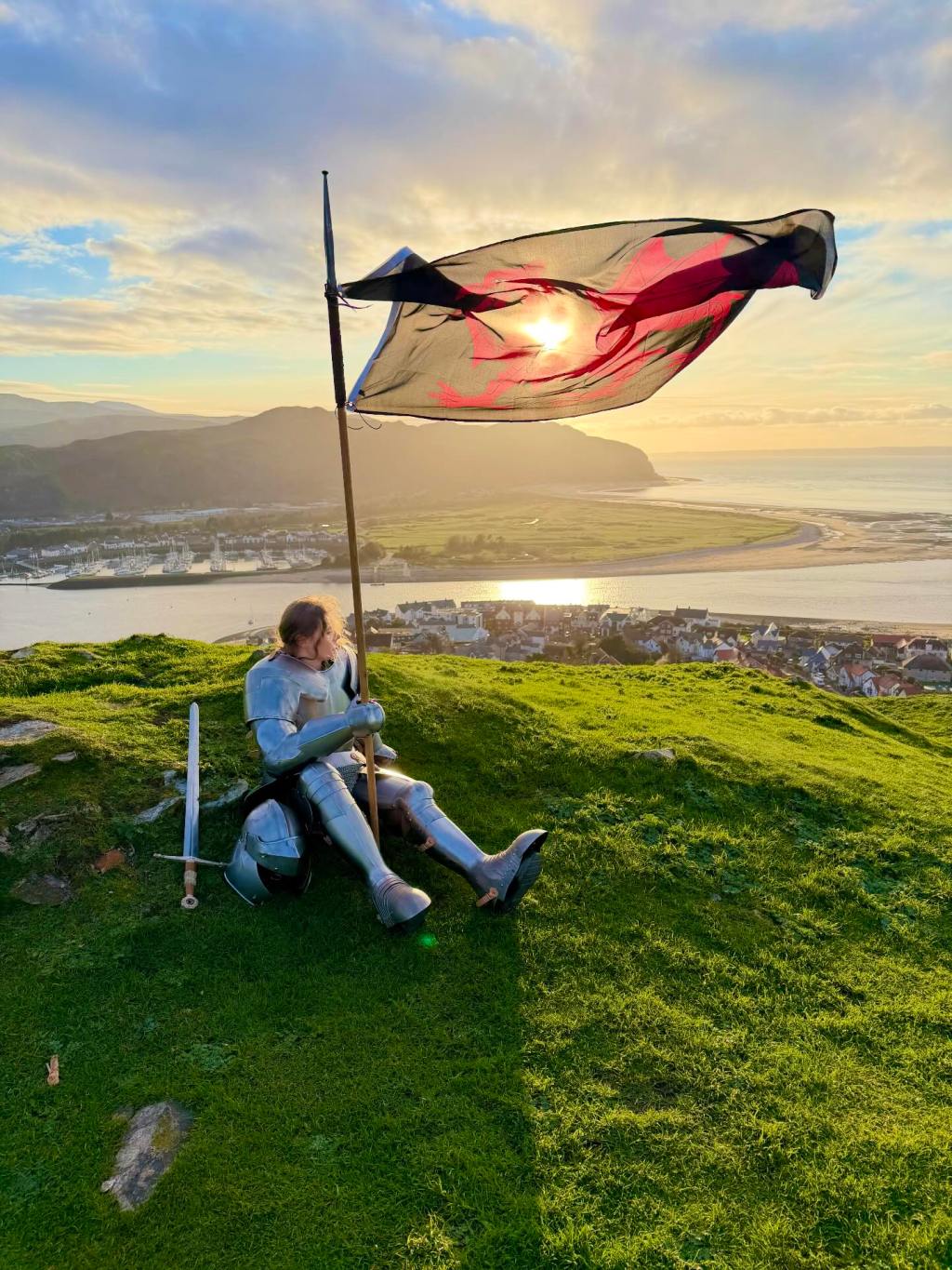 A person in shining medieval-style armor sits on a grassy hill under a dramatic sky, holding a large flagpole with a flag waving in the wind. The flag features a bright red dragon silhouetted against the setting sun, which casts a warm glow over the scene. Beside them on the grass rests a sword and helmet. The background reveals a panoramic view of a small coastal town with a river flowing into a bay, surrounded by rolling hills. The sky is mostly clear with some clouds catching the golden light of the sunset, creating a serene and picturesque atmosphere.