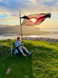 A person in shining medieval-style armor sits on a grassy hill under a dramatic sky, holding a large flagpole with a flag waving in the wind. The flag features a bright red dragon silhouetted against the setting sun, which casts a warm glow over the scene. Beside them on the grass rests a sword and helmet. The background reveals a panoramic view of a small coastal town with a river flowing into a bay, surrounded by rolling hills. The sky is mostly clear with some clouds catching the golden light of the sunset, creating a serene and picturesque atmosphere.