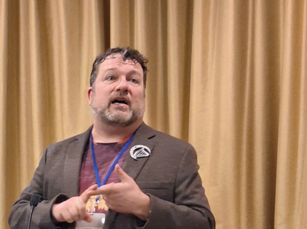 Photo of Dave Clark in a suit jacket wearing a soccer jersey while taking at an OrcaCon presentation.
